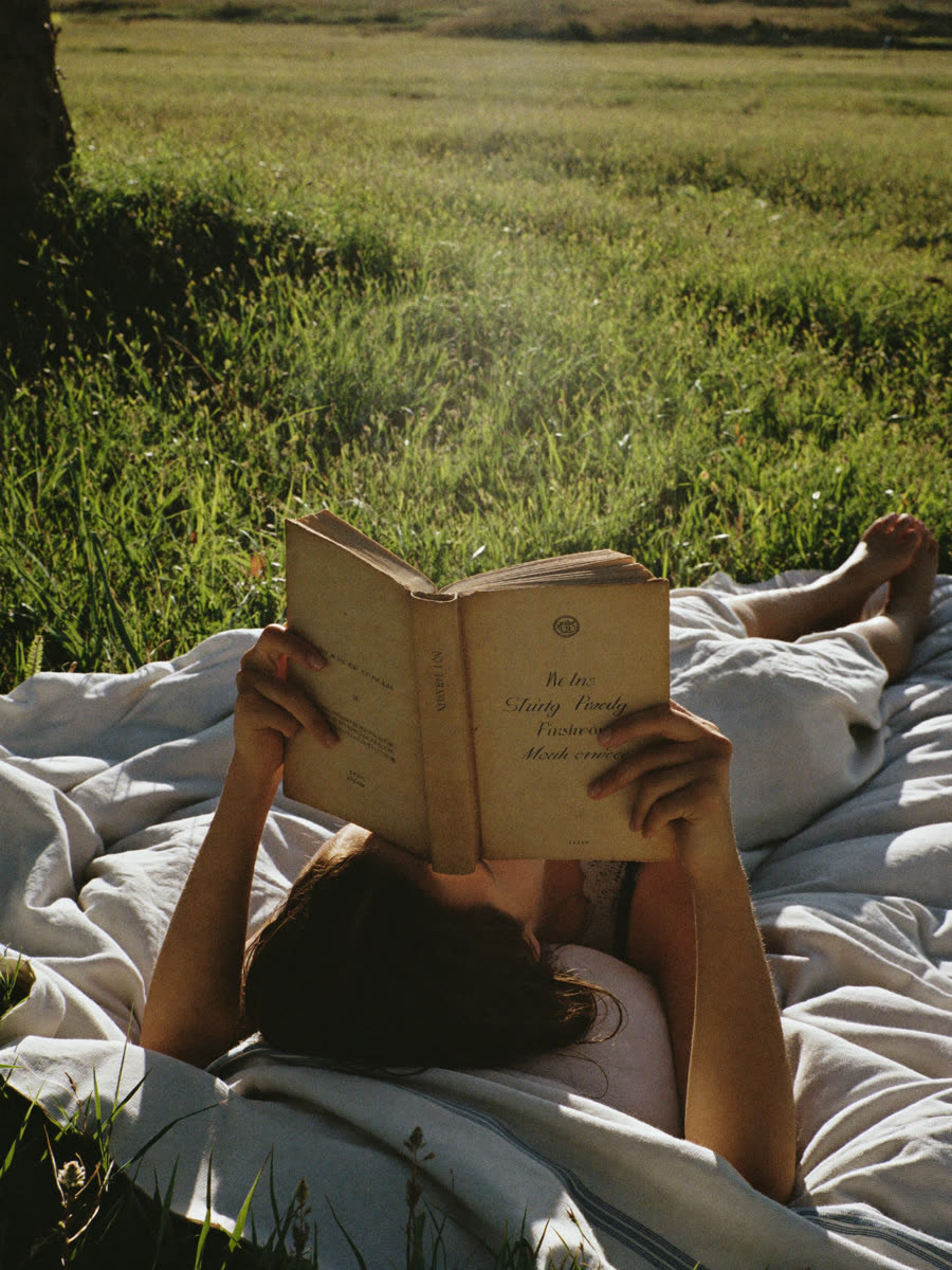 A person reading a paperback on a blanket in a sunny meadow